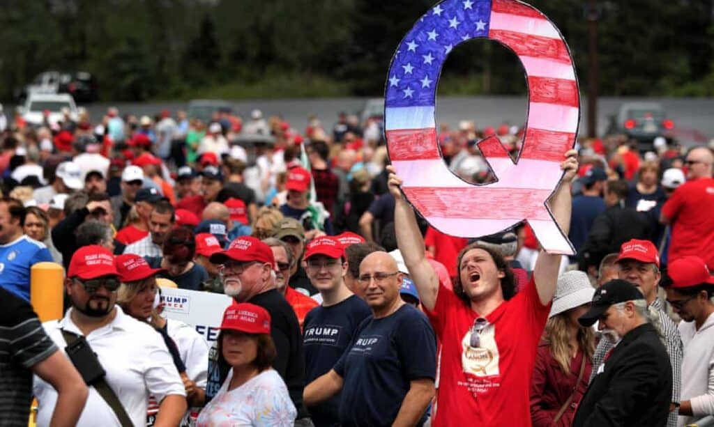 a man at a political rally holding a large cardboard cutout of a "Q," representing his support for the qanon conspiracy