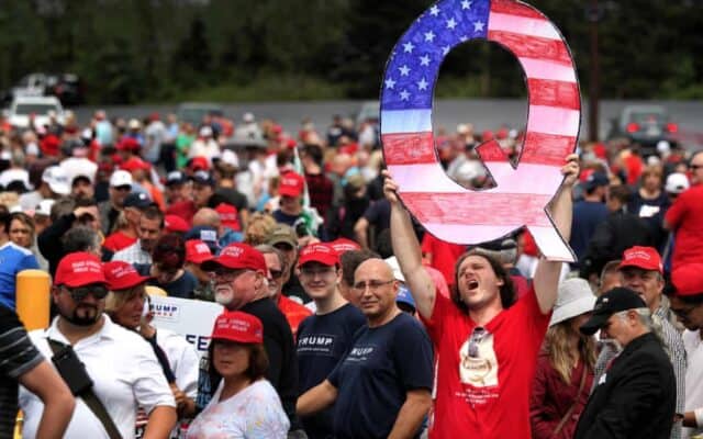 a man at a political rally holding a large cardboard cutout of a "Q," representing his support for the qanon conspiracy